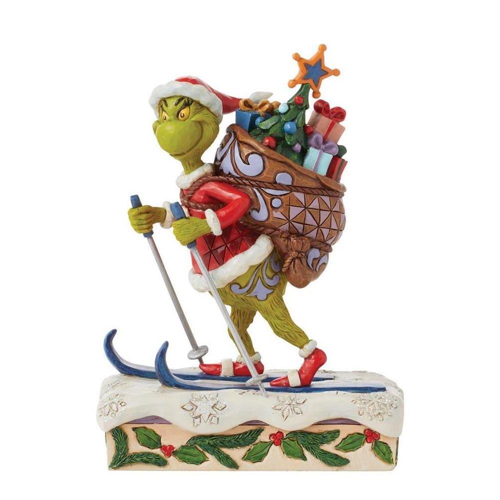 A Grinch figurine depicted skiing with poles and a large Christmas sack filled with presents and a tree, set on a snow-covered base with holly and berries, photographed on a plain white background.