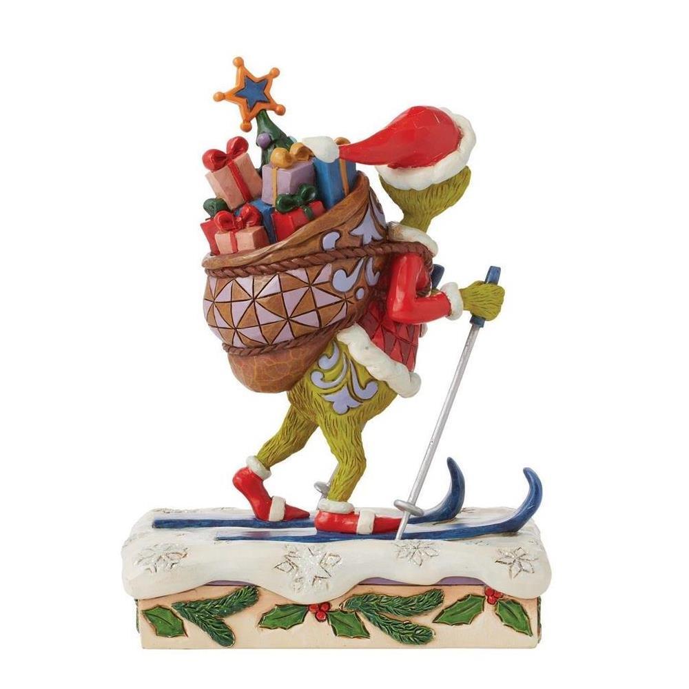 A Grinch figurine depicted skiing with poles and a large Christmas sack filled with presents and a tree, set on a snow-covered base with holly and berries, photographed on a plain white background.