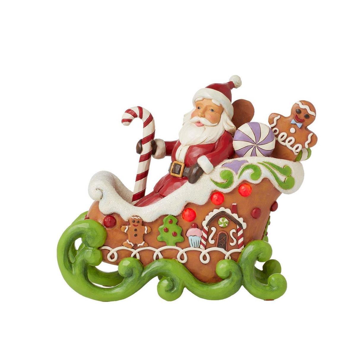 A festive Christmas ornament depicting Santa Claus riding in a gingerbread-themed sleigh, holding a candy cane, with a gingerbread man and assorted holiday decorations, on a plain white background.