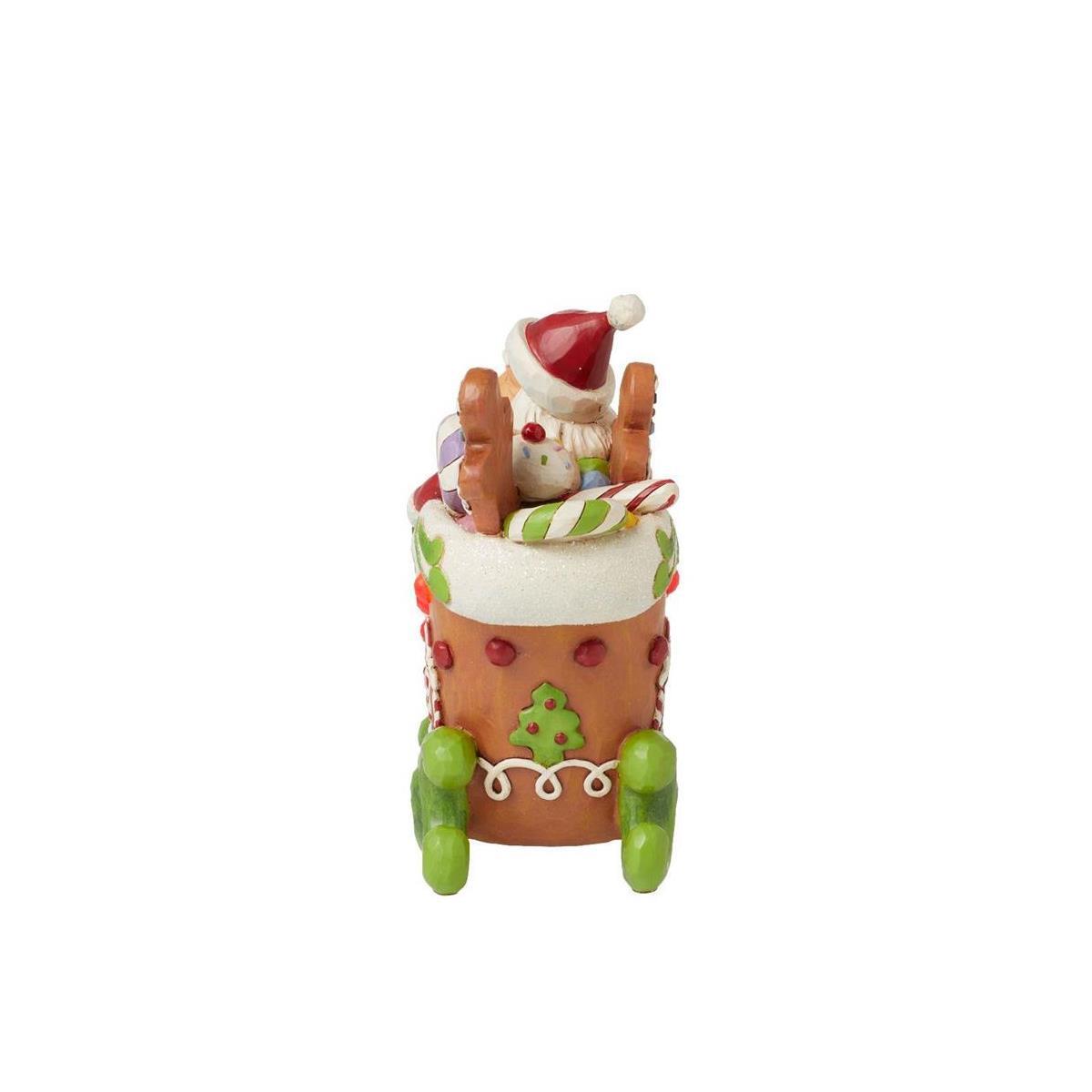 A small Christmas ornament depicting Santa Claus sitting in a gingerbread house sleigh, decorated with candy canes, green ornaments, and a white Santa hat, set against a plain white background.
