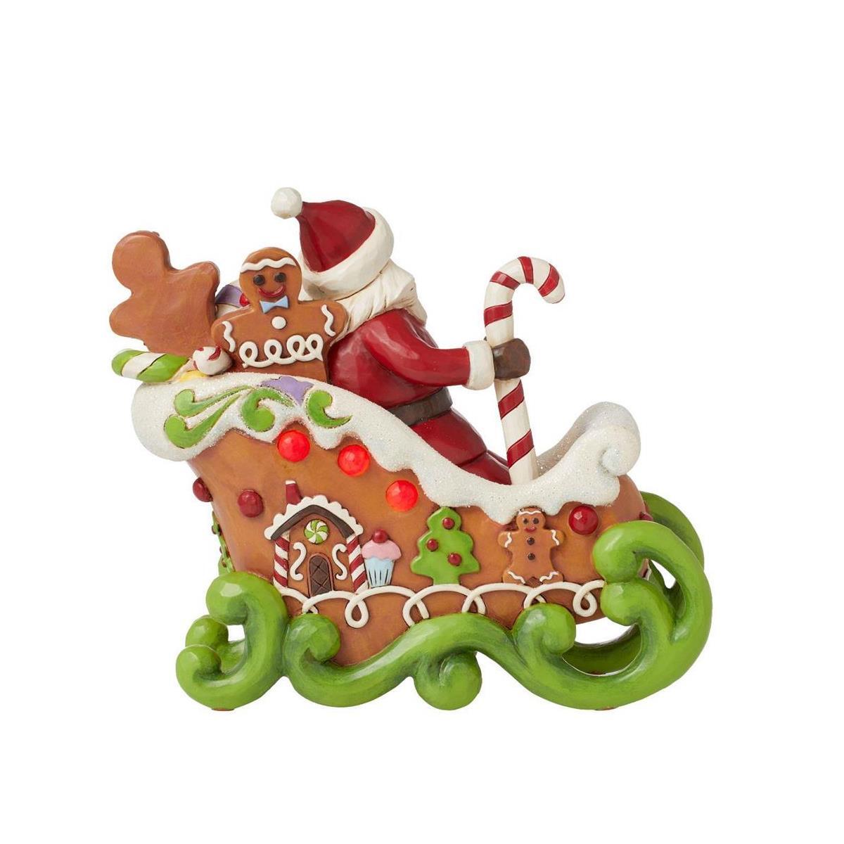 A festive Christmas ornament depicting Santa Claus in a gingerbread sleigh, holding a candy cane, with gingerbread men and houses decorated on the sleigh and a green scrollwork base, isolated on a white background.