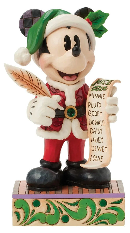 A Disney Mickey Mouse Christmas figurine wearing a Santa hat and red Santa suit, holding a feather quill and a scroll listing names, standing on a decorated base.
