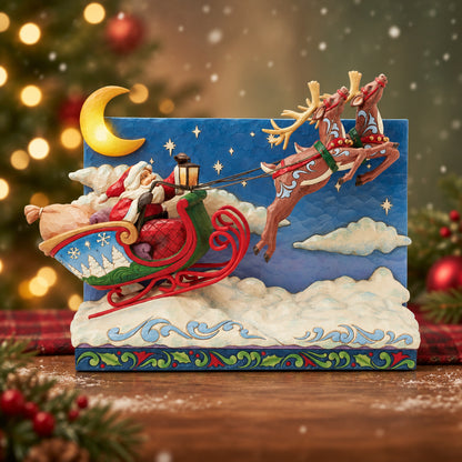 A decorative Santa Claus holiday figurine showing Santa in a red sleigh pulled by three brown reindeer against a night sky backdrop with a yellow crescent moon and clouds.