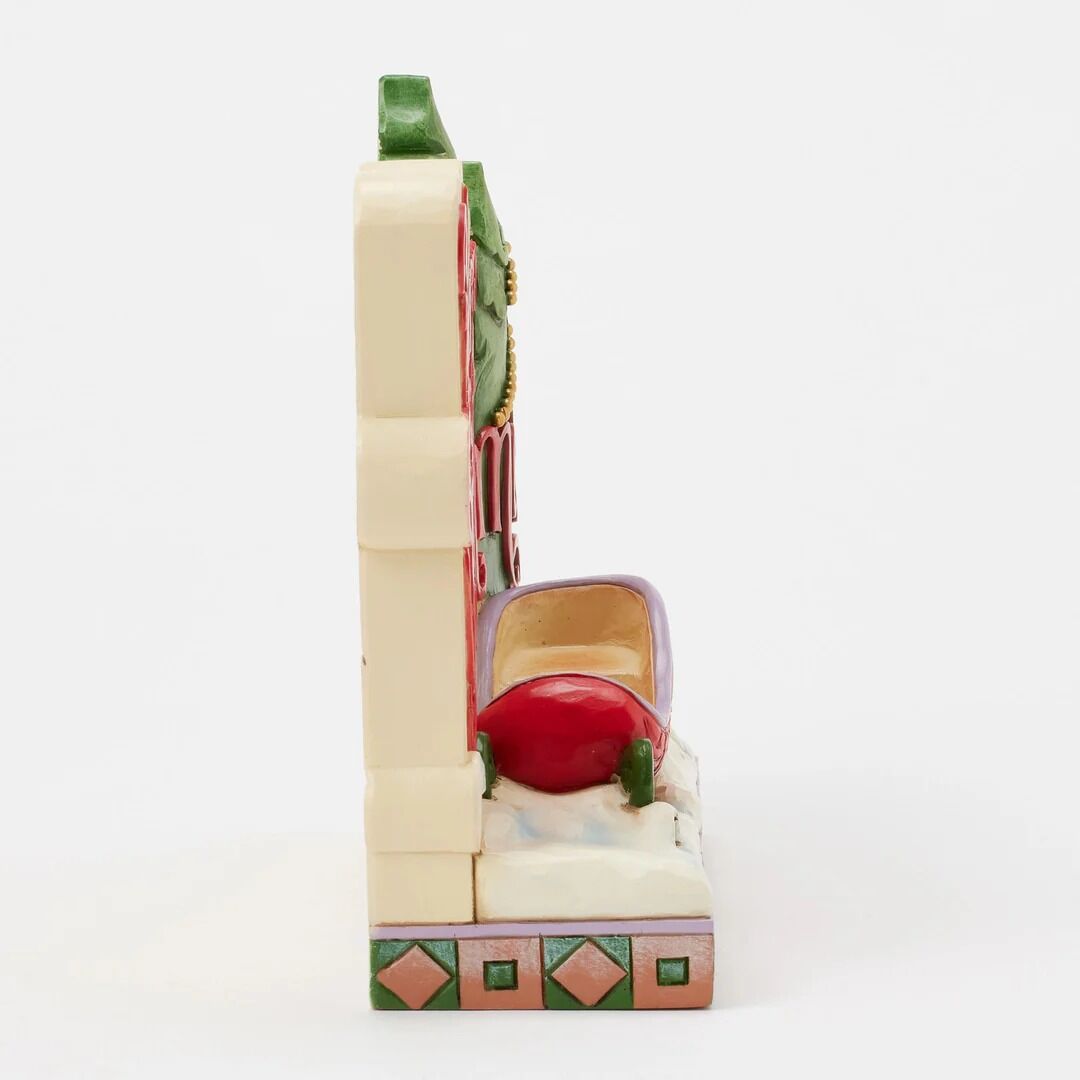 A vintage ceramic collectible figurine depicting a woman in a red dress and purple hat leaning against a cream-colored building with green and pink decorative accents, set against a plain white background.