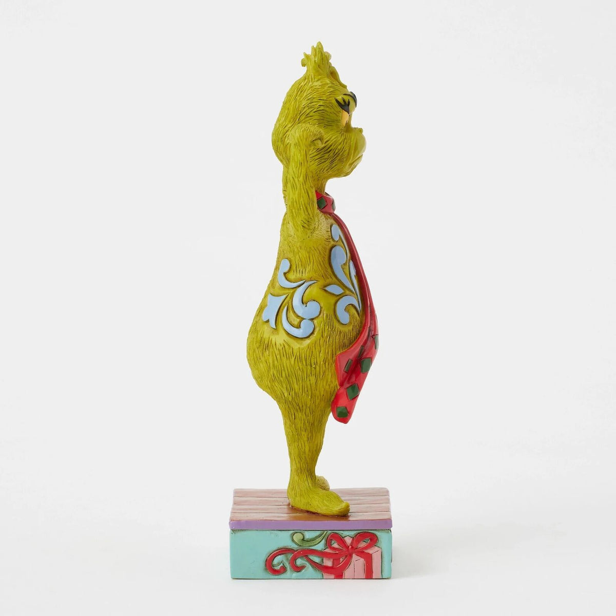 A collectible figurine of the Grinch in profile, standing on a decorated gift box. The figurine is primarily yellow-green with light blue and white swirl patterns on its back, a red and green striped cape, and stands on a pink and teal base with a red ribbon.
