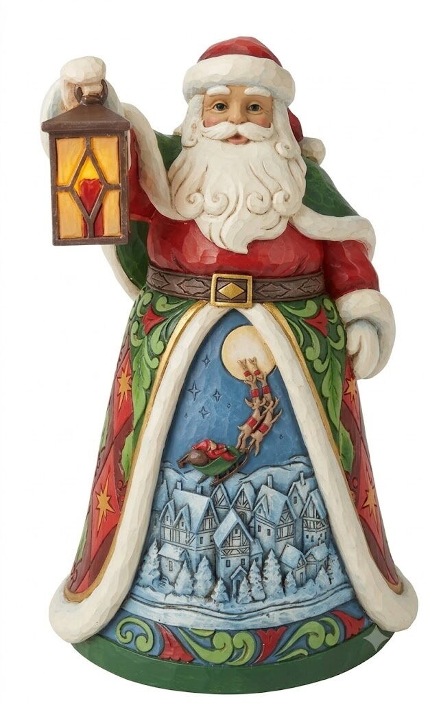 A Santa Claus figurine holding a lit lantern, featuring a detailed winter village scene on the front of his robe, set against a plain white background.