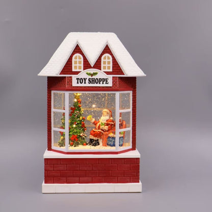 A small red and white toy house decoration with a snow-covered roof, labeled 'TOY SHOPPE' above a window scene showing Santa Claus and a Christmas tree, photographed on a plain gray background.