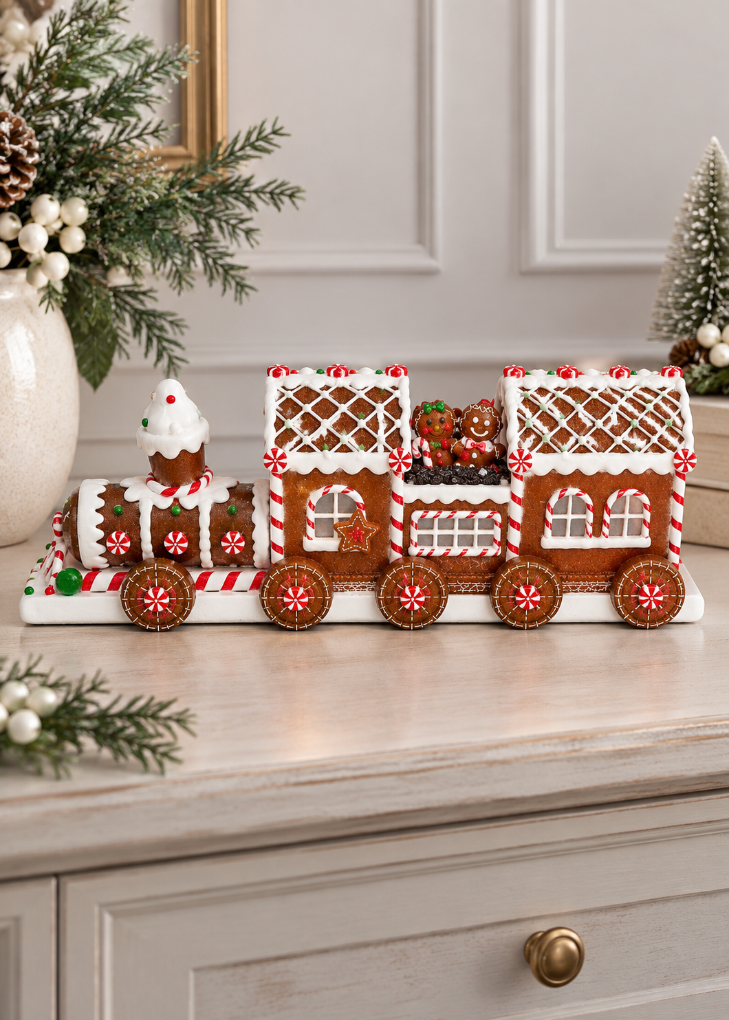 A gingerbread train decoration displayed on a light-colored wooden cabinet in a decorated holiday setting, with pine branches, a white vase, and a small Christmas tree in the background.