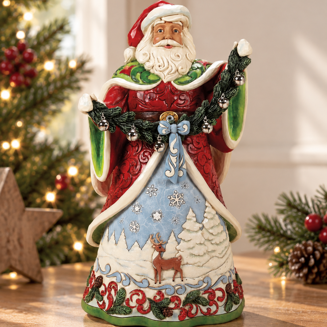 Jim Shore Heartwood Creek Santa Claus figurine standing on a wooden table with a blurred Christmas tree and holiday decor in the background. The Santa figure is decorated in red and green with a light blue skirt featuring a white reindeer and snowy trees.