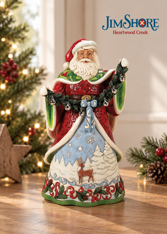 Jim Shore Heartwood Creek Santa Claus figurine standing on a wooden table with a blurred Christmas tree and holiday decor in the background. The Santa figure is decorated in red and green with a light blue skirt featuring a white reindeer and snowy trees.