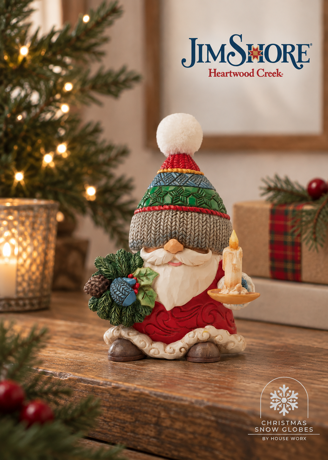 A Jim Shore Heartwood Creek Christmas snow globe figurine of a gnome in a knit hat and red coat, holding a wreath and a candle, displayed on a wooden table with a blurred Christmas tree and gift box in the background.