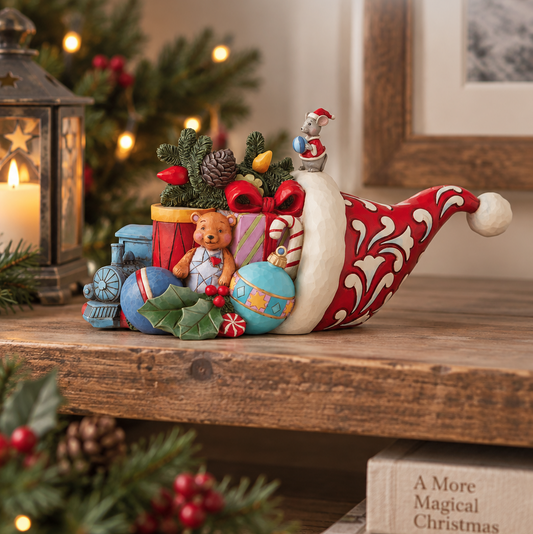 A decorative Christmas Santa hat filled with holiday gifts, ornaments, a teddy bear, pinecones, and small figurines, displayed on a wooden shelf beside a lantern and Christmas tree. Visible text includes 'Christmas Snow Globes by House Work' and 'Jim Shore Heartwood Creek'.