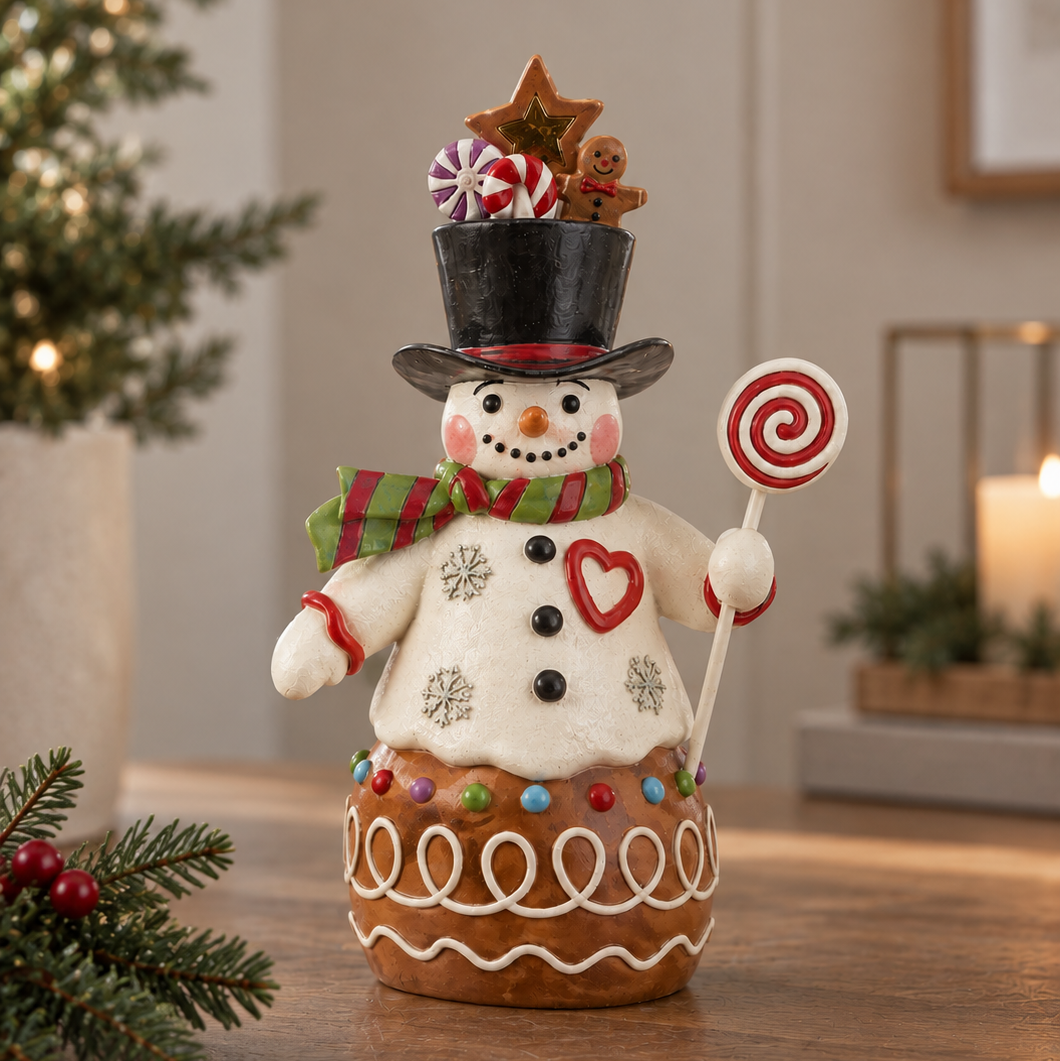 A festive Christmas snowman figurine standing on a decorated gingerbread base, placed on a wooden table in a warmly lit indoor setting with holiday greenery and candles in the background.