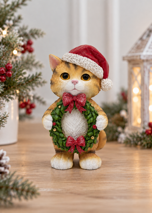 A small ginger cat figurine wearing a red Santa hat and a green holiday wreath with red bows, standing on a wooden surface in a softly lit Christmas setting with pine branches and a lantern in the background.