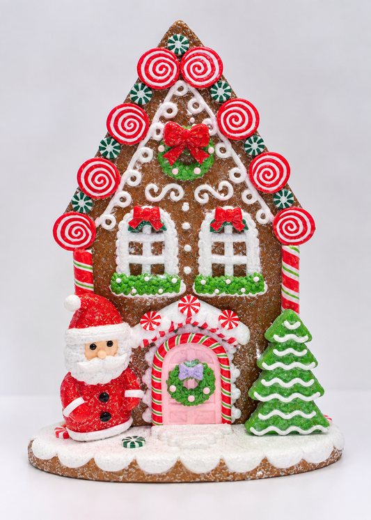 A festive gingerbread house decoration decorated with white icing, red and green candy swirls, a red bow, a pink door, a green tree, and a small Santa Claus figure, set against a plain light gray background.