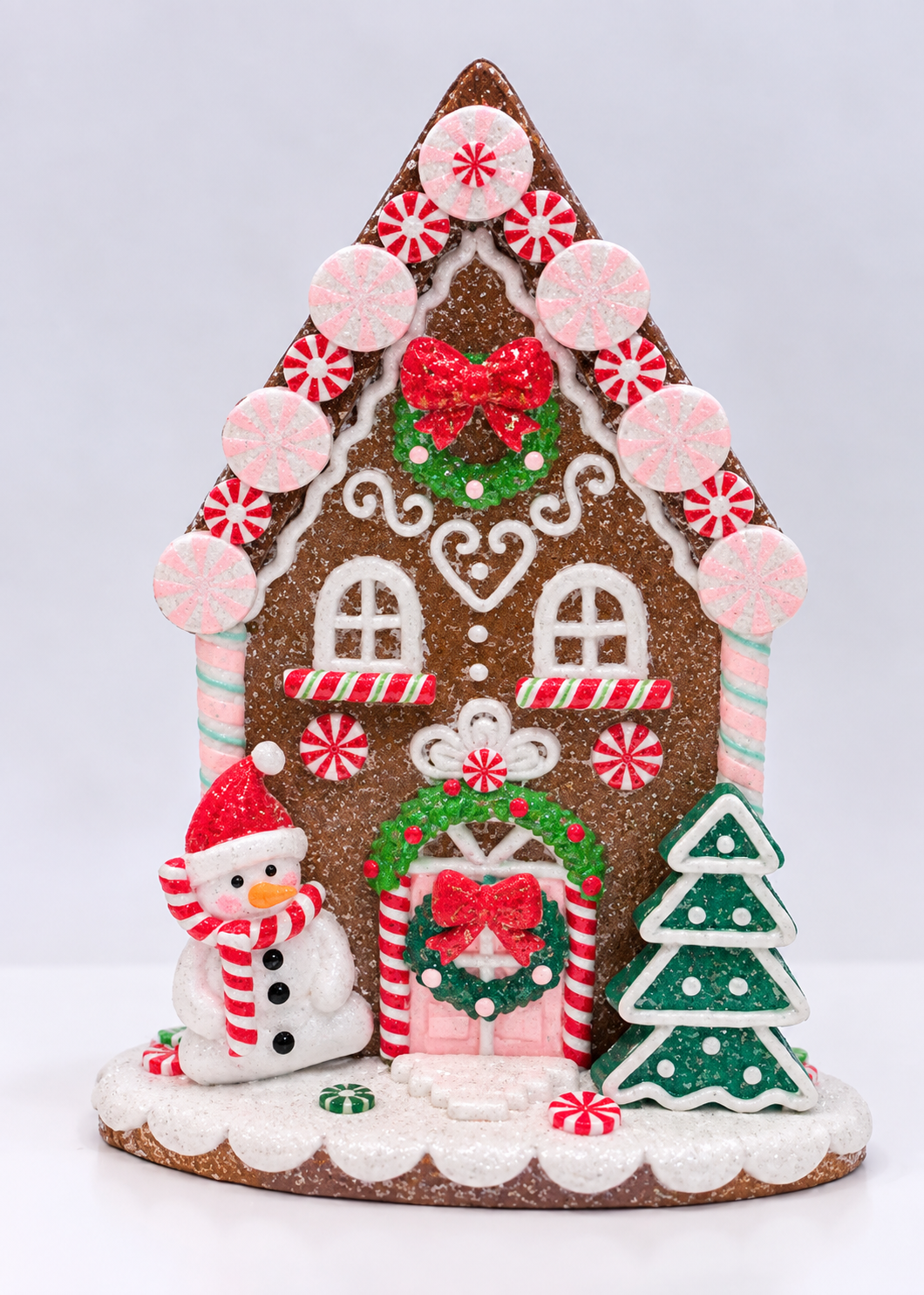 A festive gingerbread house Christmas decoration with white icing details, pink candy canes, a green wreath with red bows, a snowman, and a green Christmas tree on a white background.