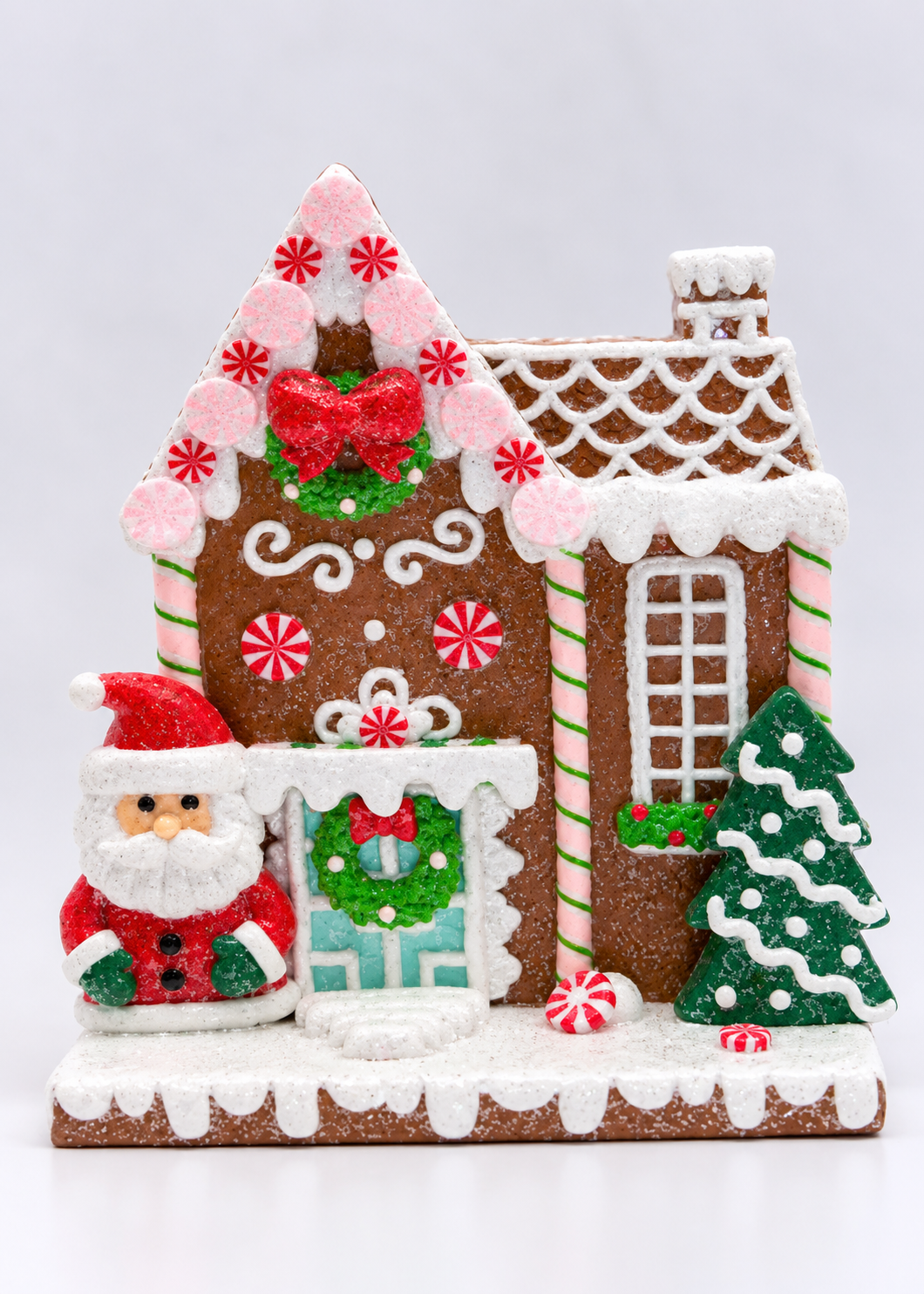 A festive gingerbread house decoration with white icing-style snow, candy canes, a green wreath, a red bow, a Santa Claus figure, and a small decorated Christmas tree on a white studio background.