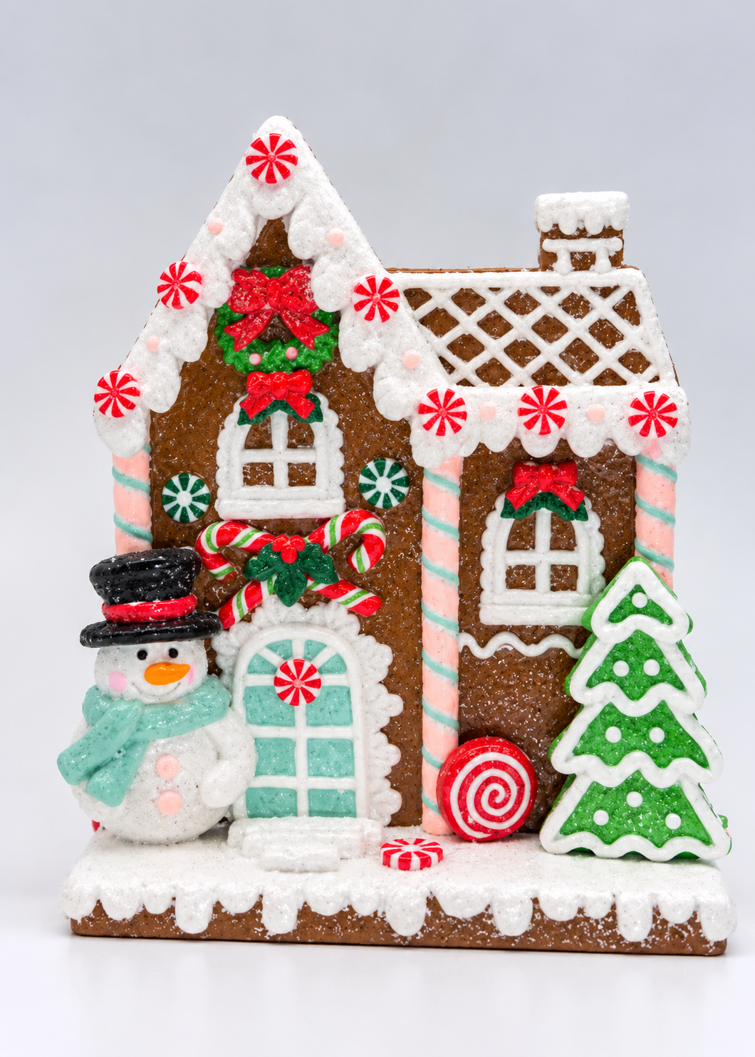 A festive gingerbread house decoration with a snowman, candy tree, and red and green holiday accents, photographed on a clean white background.