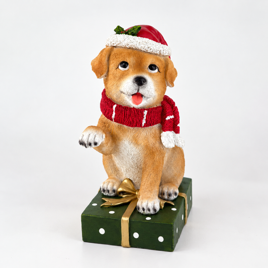 A decorative figurine of a smiling tan puppy wearing a red knit Santa hat and matching red knit scarf, sitting on a dark green gift box with white polka dots and a gold ribbon, photographed on a clean white studio background.