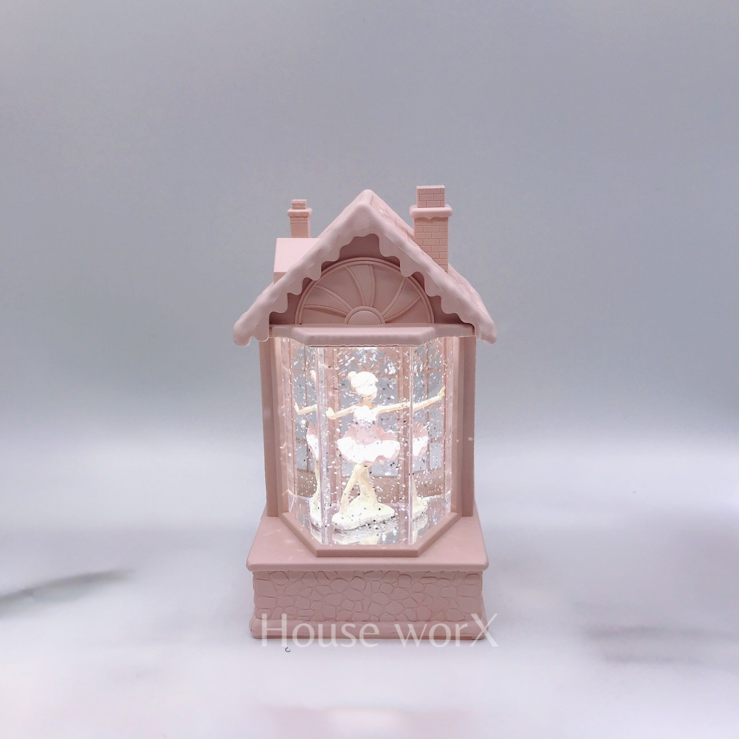 A small pink house-shaped snow globe figurine with a ballerina inside, photographed against a soft gray studio background.
