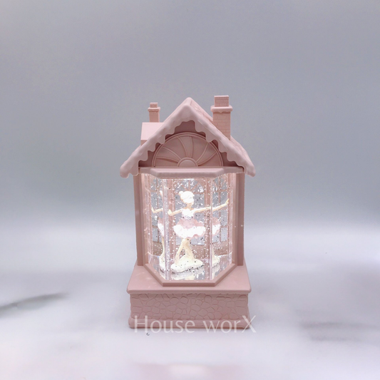 A small pink house-shaped snow globe figurine with a ballerina inside, photographed against a soft gray studio background.