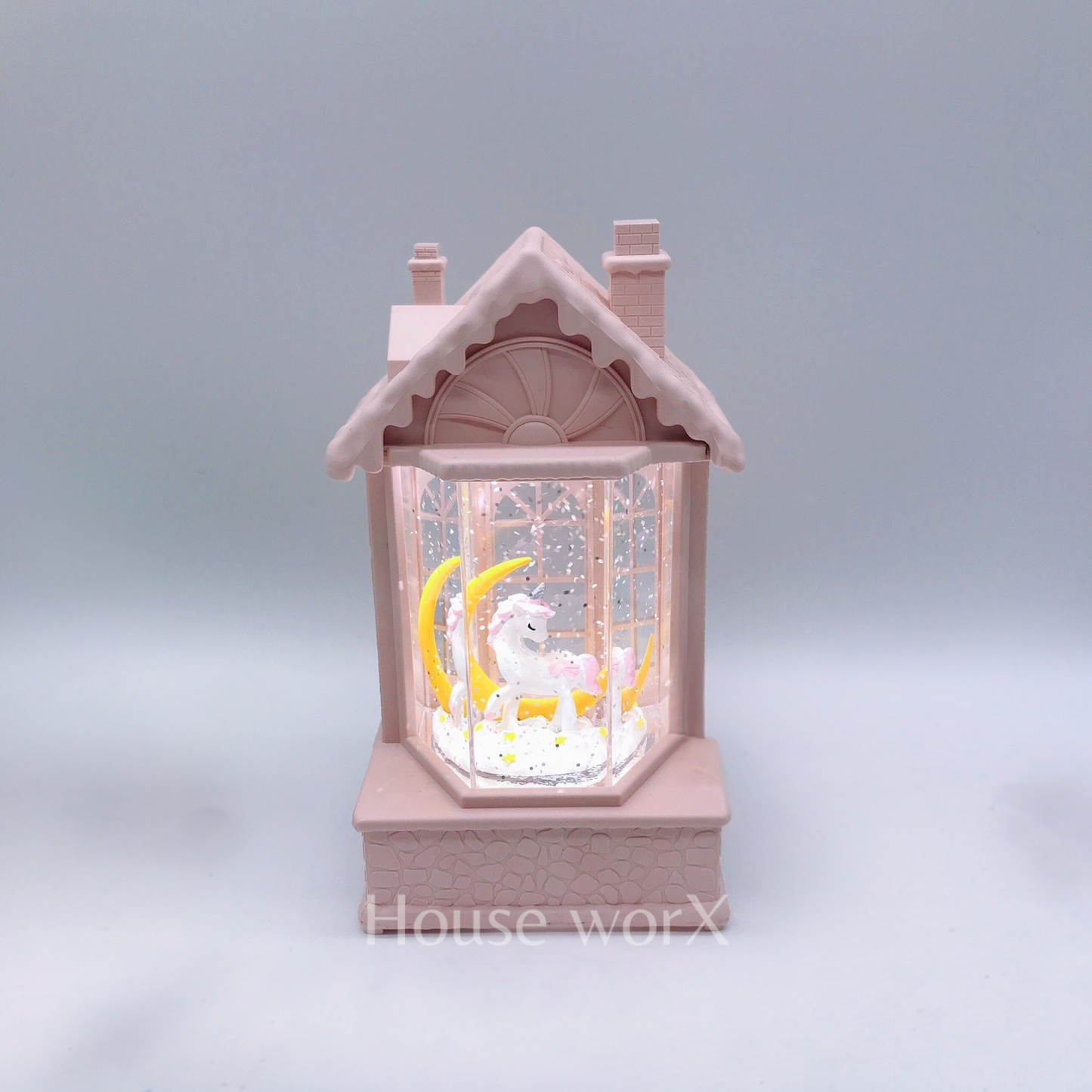 A pink house-shaped snow globe night light featuring a white unicorn and yellow crescent moon inside, set against a soft gray background.