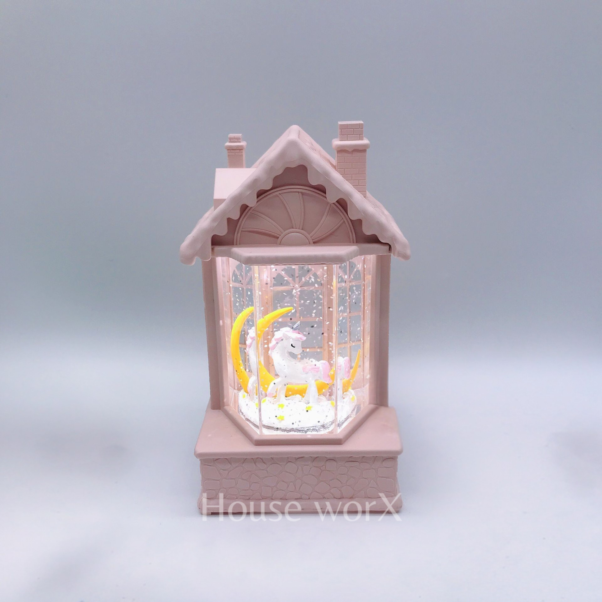 A pink house-shaped snow globe night light featuring a white unicorn and yellow crescent moon inside, set against a soft gray background.
