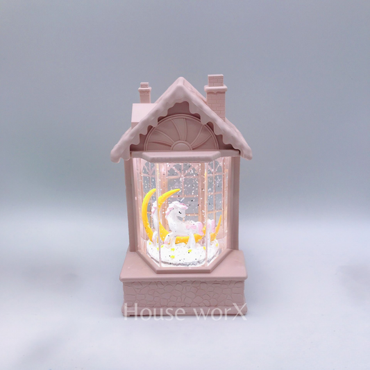 A pink house-shaped snow globe night light featuring a white unicorn and yellow crescent moon inside, set against a soft gray background.