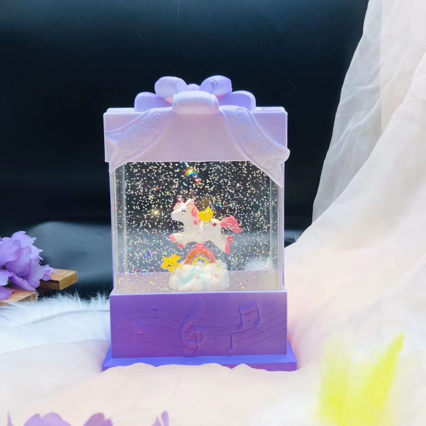A purple gift-box-shaped snow globe music box with a unicorn and rainbow design inside, set against a soft white fabric backdrop with a dark background.