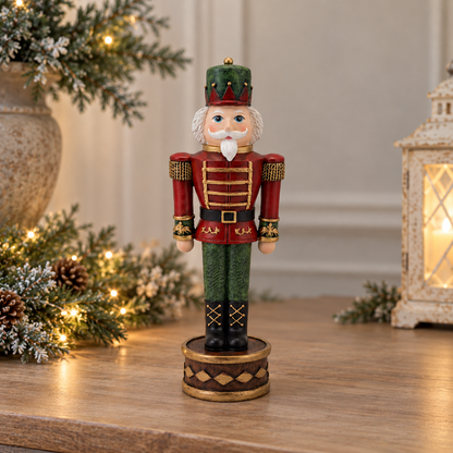 A decorative Christmas nutcracker figurine standing on a round base, placed on a wooden surface with holiday greenery and a lantern in the softly blurred background.
