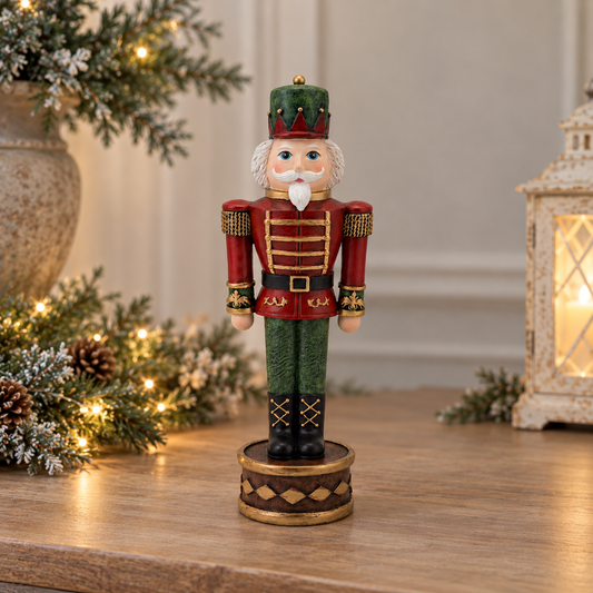 A decorative Christmas nutcracker figurine standing on a round base, placed on a wooden surface with holiday greenery and a lantern in the softly blurred background.