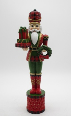 A festive nutcracker figurine wearing a red and green plaid uniform, holding a wreath and gift boxes, standing on a red patterned cylindrical base against a light gray background.