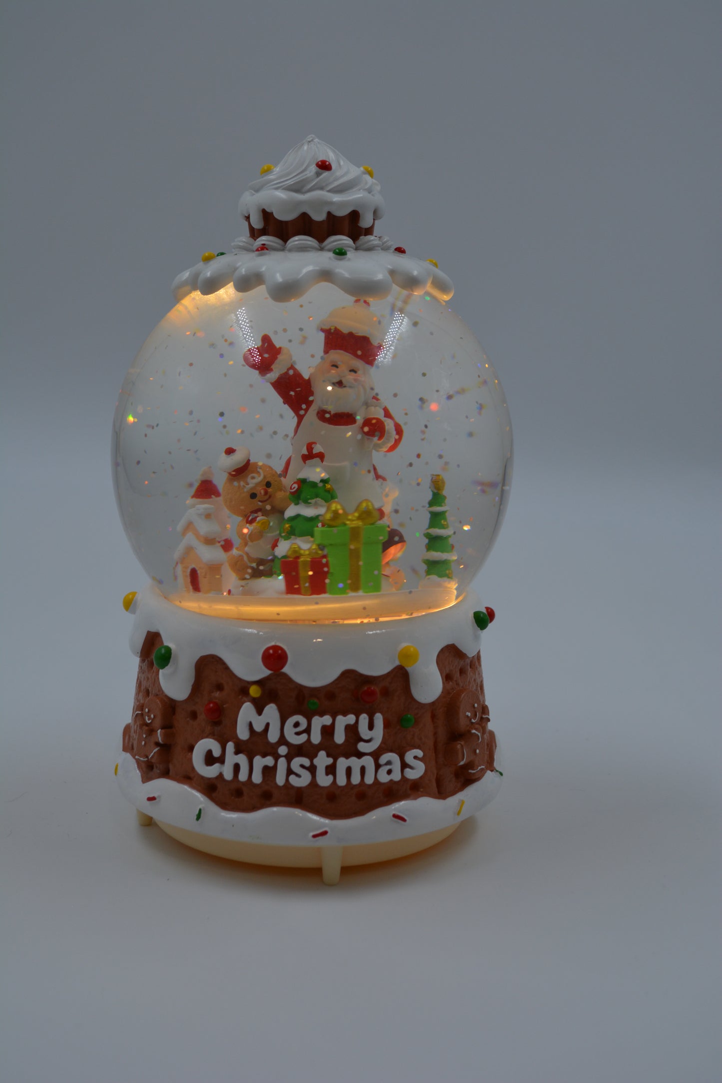 A festive Christmas snow globe figurine shaped like a gingerbread cake, featuring a Santa Claus and teddy bear scene inside, with 'Merry Christmas' written on the front, set against a neutral gray background.