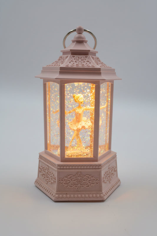 A pink decorative lantern with a ballerina figurine inside, illuminated with a warm light against a neutral gray background.