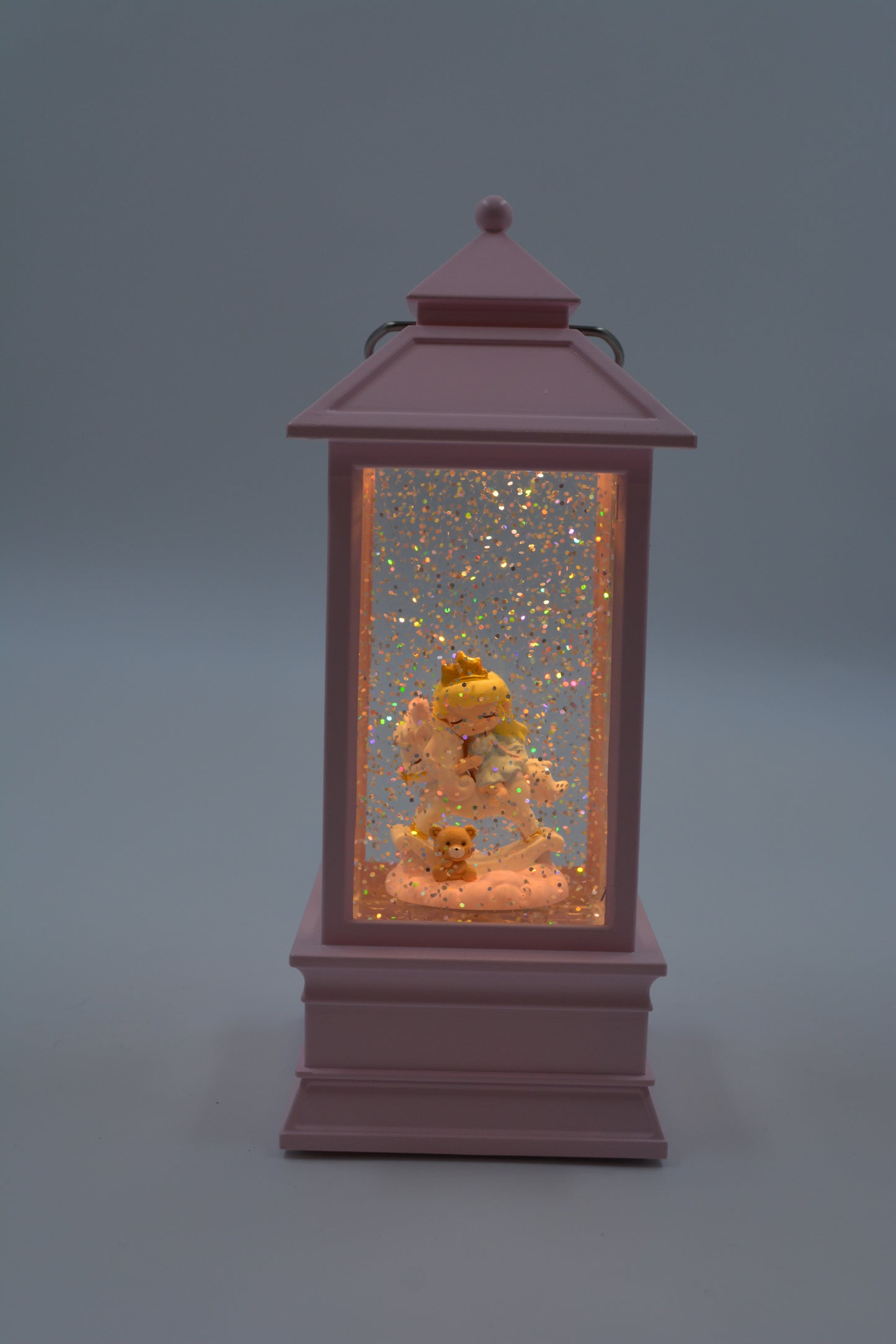 A pink lantern-shaped decorative snow globe with a warm internal light, featuring a small angel figurine surrounded by glittery gold flakes and white faux snow on a soft gray background.