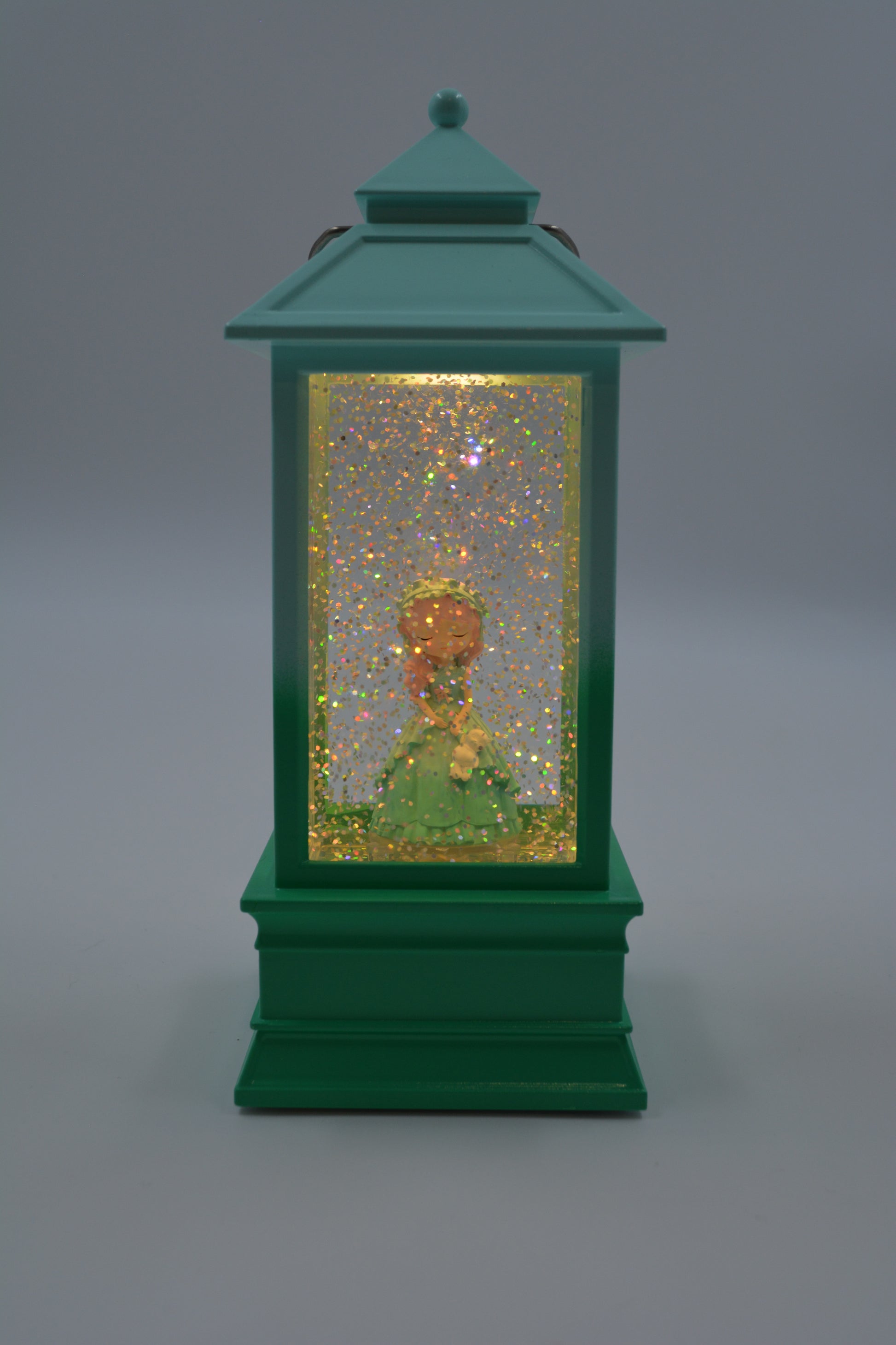 A green lantern-shaped decorative object with a glitter-filled interior and a small gold-and-green figurine inside, photographed against a smooth gray studio background.