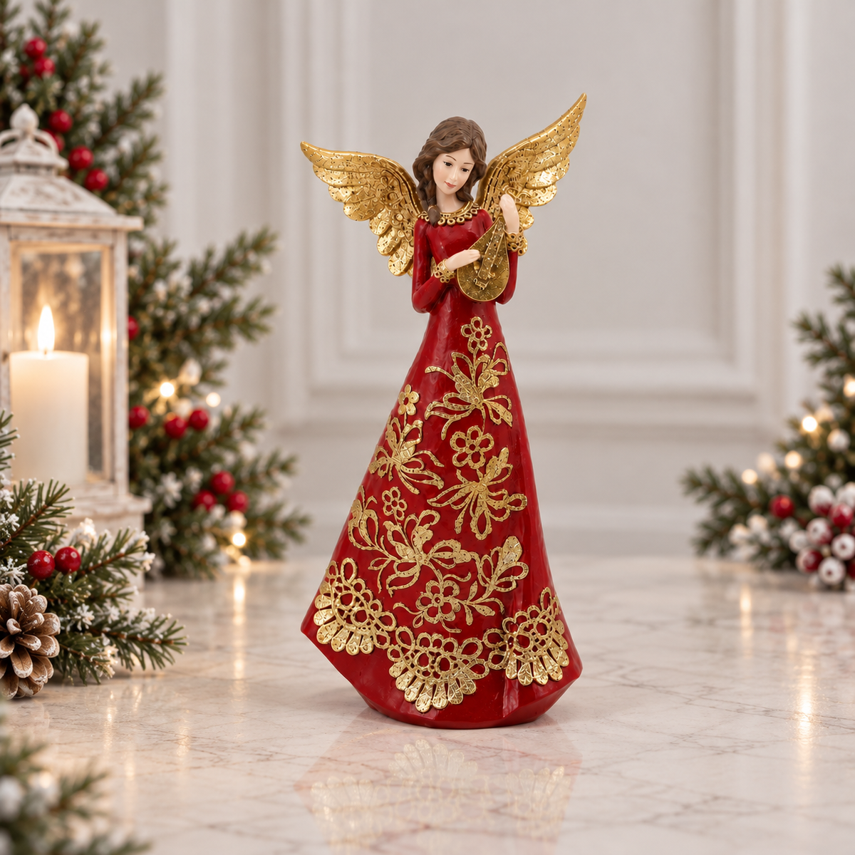 A Christmas angel figurine wearing a long red gown with gold floral embroidery and gold wings, standing on a light marble surface with holiday greenery, a lantern candle, and warm lights in the background.