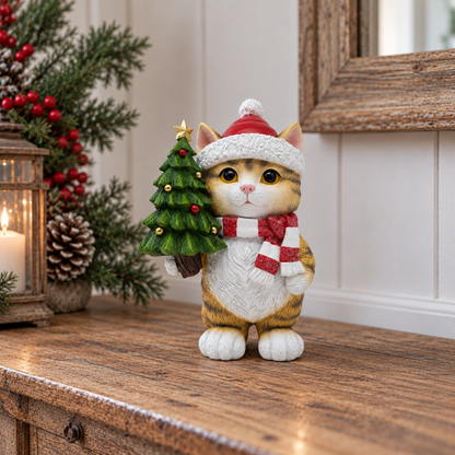 A festive cat figurine wearing a white Santa hat and red-and-white striped scarf, holding a small green Christmas tree, displayed on a wooden surface in a holiday-themed setting.