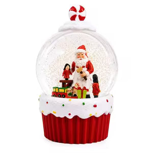 A festive Christmas snow globe figurine shaped like a cupcake, featuring a red base with white frosting-like trim, a clear dome containing Santa Claus, a penguin, a small train, and gift boxes, topped with a candy cane ornament.