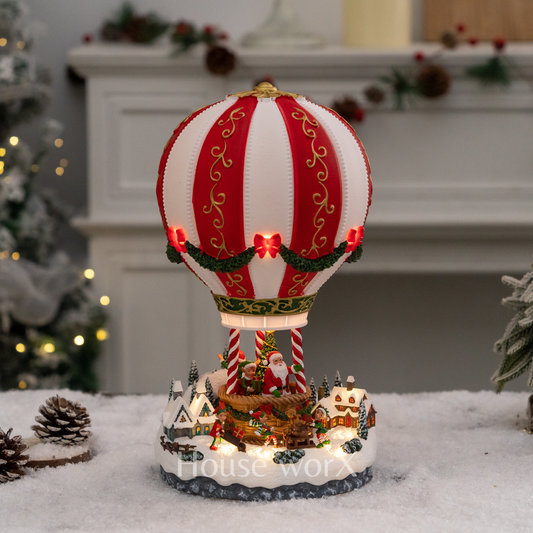 Decorative hot air balloon with Santa Claus on a snowy base, Christmas tree and fireplace in the background.