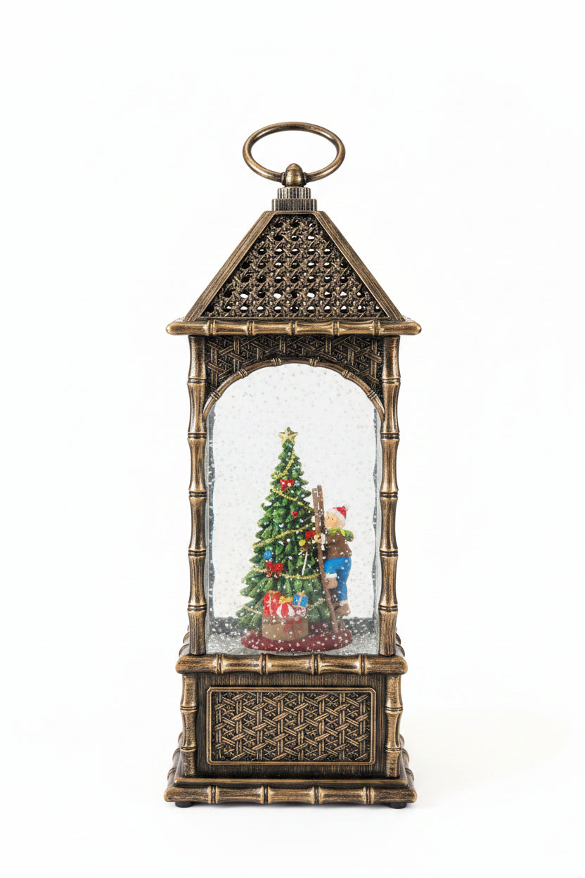 A decorative lantern-shaped snow globe with a gold and bronze bamboo-style frame, featuring a miniature Christmas tree scene inside, photographed on a kitchen countertop with a white sheet of paper behind it.