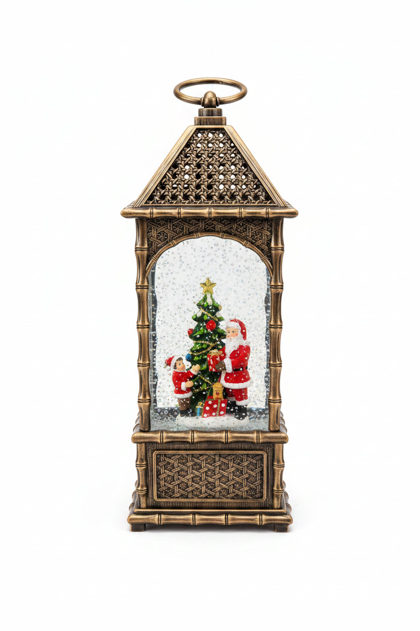 A decorative lantern-shaped Christmas ornament with a gold-toned metal frame, featuring a miniature Christmas tree, Santa Claus figures, and white faux snow inside, displayed on a kitchen countertop with a white sheet of paper behind it.