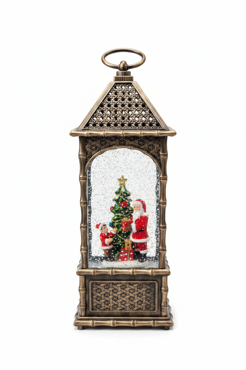 A decorative lantern-shaped Christmas ornament with a gold-toned metal frame, featuring a miniature Christmas tree, Santa Claus figures, and white faux snow inside, 