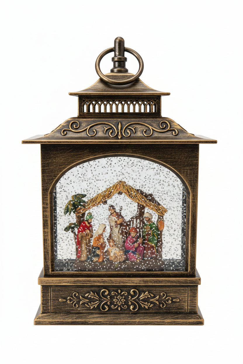 An ornate bronze-finished lantern-shaped Christmas nativity scene with a glass window, internal stable structure, and small figurines under a faux snow effect, shown on a countertop.