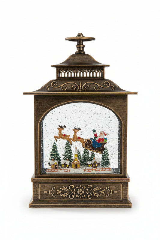 A decorative tabletop lantern-style Christmas ornament with an antique bronze finish and a snow globe scene inside featuring Santa Claus, reindeer, trees, and small houses, shown on a granite countertop.