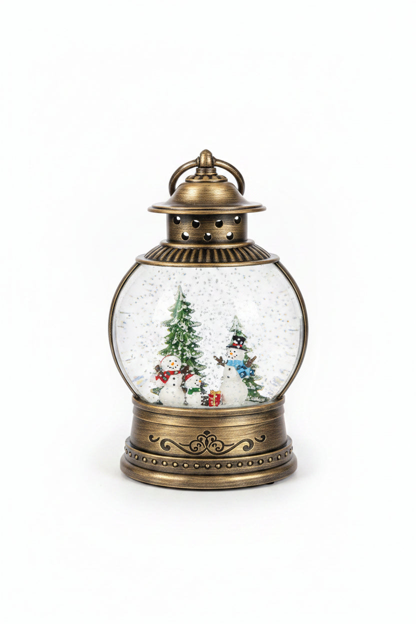 A vintage-style holiday snow globe lantern with an antique bronze metal frame, featuring a miniature winter scene inside with snowmen, a Christmas tree, and falling glittery snow, displayed on a granite countertop.