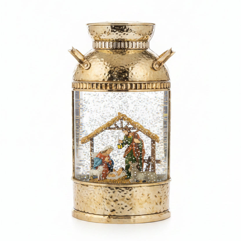 A decorative snow globe lantern shaped like a vintage galvanized metal milk canister, featuring a gold-toned hammered finish, side handles, and a clear glass body filled with falling snow and a nativity scene figurine inside.