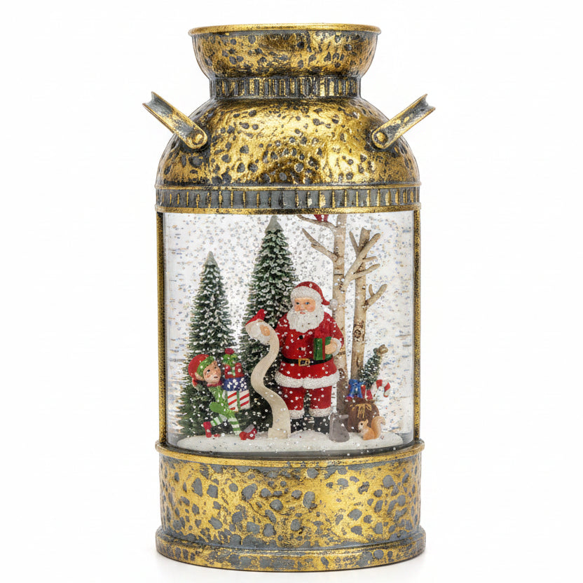 A decorative vintage-style milk canister snow globe with a hammered gold and silver finish, featuring Santa Claus, a reindeer, and Christmas trees inside a clear glass chamber, photographed on a kitchen countertop.