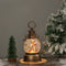 A vintage style camp lantern with a nativity design, featuring a bronze-colored base and a clear globe with a nativity scene inside, placed on a wooden surface with snow and a Christmas tree in the background.