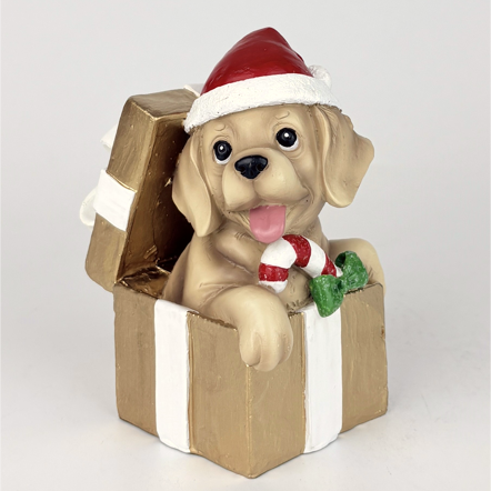 A decorative figurine of a light tan puppy wearing a red and white Santa hat and holding a red-and-white candy cane, sitting inside an open gold gift box with white ribbon and green holly accents, on a light gray background.